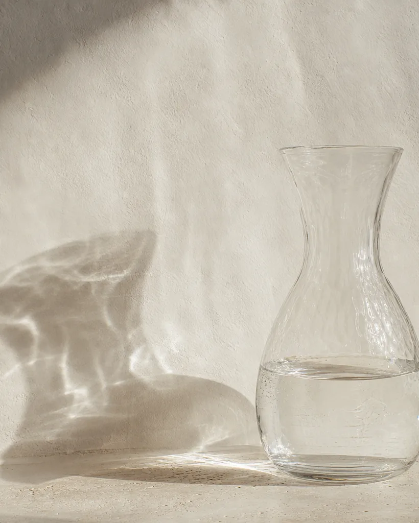 A hand-blown clear glass carafe refracting sunlight onto a pale wall.